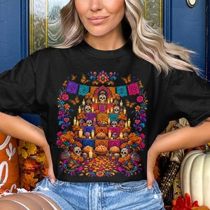 May include: A black t-shirt featuring a vibrant graphic print of a Day of the Dead altar. The design includes colorful skulls, marigold flowers, lit candles, butterflies, and decorative papel picado banners arranged in a tiered display.