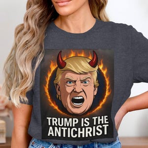 May include: Dark gray t-shirt featuring a graphic of a man with blonde hair and red horns, surrounded by flames. The text "TRUMP IS THE ANTICHRIST" is printed below the image.