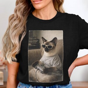 May include: Black t-shirt featuring a sepia-toned image of a Siamese cat wearing sunglasses and a t-shirt, holding a video game controller. The cat is seated on a couch. The image has a vintage aesthetic.