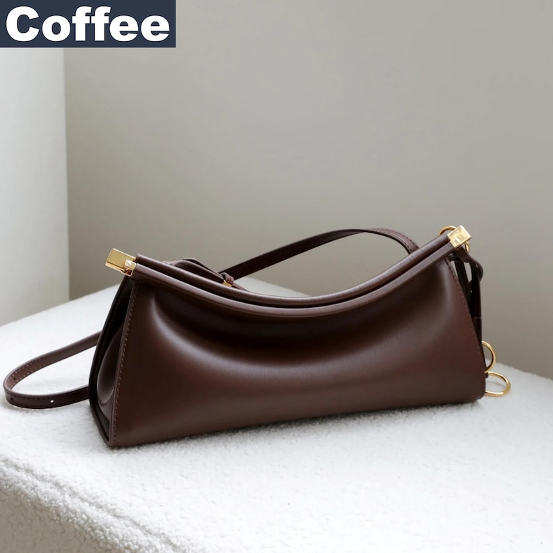 May include: A brown leather handbag with gold-colored hardware. The bag has a curved top handle and a long, adjustable shoulder strap. The word "Coffee" is in a white rectangle in the upper left corner.