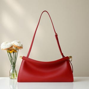 May include: A vibrant red handbag with a curved top handle and a long, adjustable shoulder strap. The bag features gold-toned hardware and a sleek, minimalist design. A small glass vase with white and yellow flowers is to the left.