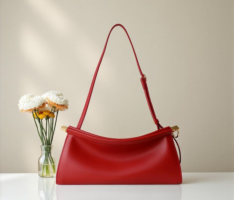 May include: A vibrant red handbag with a curved top handle and a long, adjustable shoulder strap. The bag features gold-toned hardware and a sleek, minimalist design. A small glass vase with white and yellow flowers is to the left.