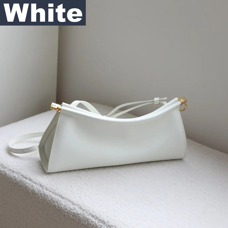 May include: A white handbag with a curved top and a long, thin strap. The bag has gold-colored hardware and the word "White" in a dark blue box in the upper left corner. The bag is sitting on a white surface.
