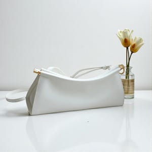 May include: A white leather handbag with a curved design and a long, thin strap. The bag has gold-colored hardware and a minimalist aesthetic. A small vase with two flowers is in the background.