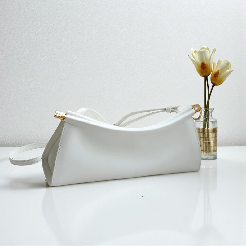 May include: A white leather handbag with a curved design and a long, thin strap. The bag has gold-colored hardware and a minimalist aesthetic. A small vase with two flowers is in the background.