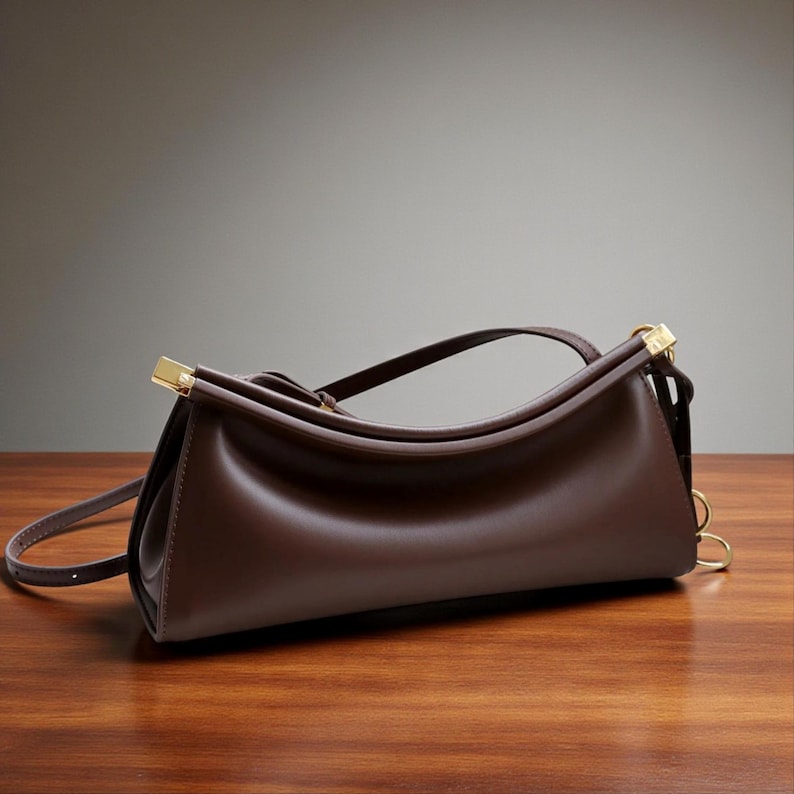 May include: A dark brown leather handbag with a curved top handle and a long, adjustable shoulder strap. The bag features gold-colored metal accents on the handle and strap attachments. The bag is sitting on a wooden surface.