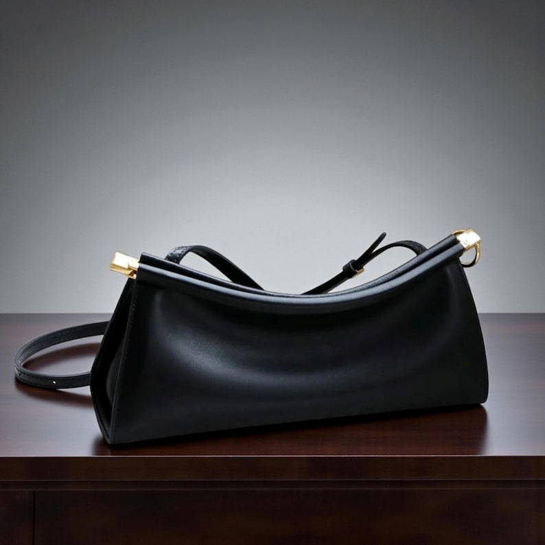 May include: A black leather handbag with a rectangular shape and a curved top. The bag features gold-colored metal accents on the handles and a long, adjustable strap. The bag is sitting on a dark brown wooden surface.