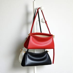 May include: Three leather handbags in red, black, and white hang from a white rack. The red bag is at the top, with a long strap. The black bag is below it, and the white bag is partially visible. All bags have gold-colored hardware.