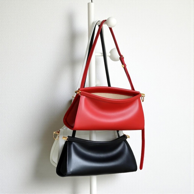 May include: Three leather handbags in red, black, and white hang from a white rack. The red bag is at the top, with a long strap. The black bag is below it, and the white bag is partially visible. All bags have gold-colored hardware.