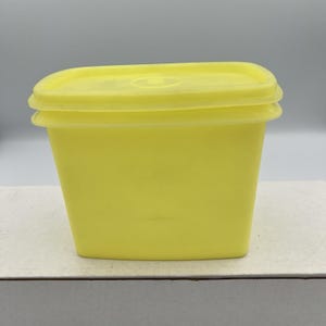 May include: A yellow plastic container with a matching lid. The container is rectangular with slightly curved sides and a flat top. The lid has a raised edge and a circular indentation in the center. The container is sitting on a white surface.