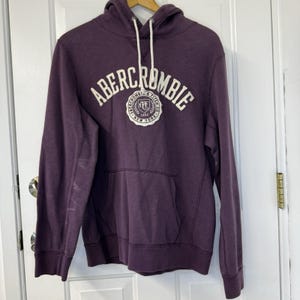 May include: A dark purple Abercrombie & Fitch hoodie with a drawstring hood and a kangaroo pocket. The brand name and logo are printed in white across the chest. The hoodie is hanging on a wooden hanger.