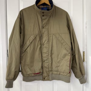 May include: A tan bomber jacket with a navy blue collar and a ribbed hem and cuffs. The jacket has a button-up front, two front pockets, and a red lining visible at the bottom.