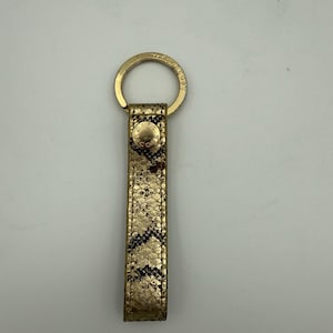 May include: Gold-coloured metal keyring with a rectangular, textured, gold and brown leather strap. The strap features a repeating chevron pattern. The keyring has a circular clasp and a small, round button with the brand name.