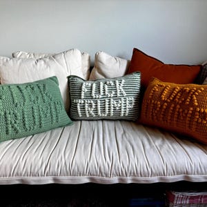 Peut inclure: Plusieurs coussins crochetés faits à la main de différentes couleurs et motifs. Un coussin vert porte l'inscription "FUCK TRUMP", un autre gris et blanc dit "FUCK TRUMP", et un troisième coussin orange dit également "FUCK TRUMP". Les coussins sont disposés sur un canapé blanc.