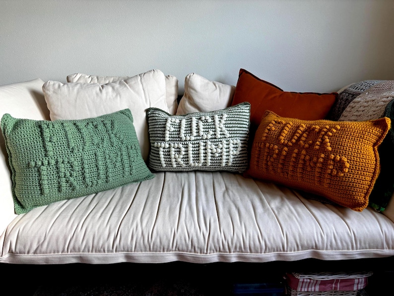 Op de afbeelding: Een verzameling handgemaakte gehaakte kussens in verschillende kleuren en ontwerpen. E&eacute;n groen kussen leest "FUCK TRUMP", een ander grijs en wit kussen zegt "FUCK TRUMP", en een derde oranje kussen zegt ook "FUCK TRUMP". De kussens zijn gerangschikt op een witte bank.