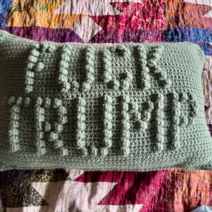 Op de afbeelding: Een rechthoekig, lichtgroen gehaakt kussen met de woorden "FUCK TRUMP" in verhoogde, getextureerde steken. Het kussen ligt op een kleurrijke, patroonquil met verschillende geometrische vormen en bloemmotieven.