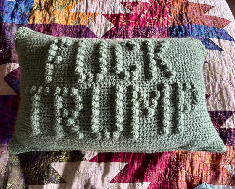 Op de afbeelding: Een rechthoekig, lichtgroen gehaakt kussen met de woorden "FUCK TRUMP" in verhoogde, getextureerde steken. Het kussen ligt op een kleurrijke, patroonquil met verschillende geometrische vormen en bloemmotieven.