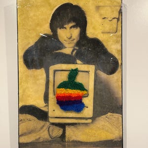 Op de afbeelding: Ingelijst kunstwerk met een persoon met een computer, met een regenboogkleurig Apple-logo. De afbeelding is in zwart-wit met een gele achtergrond. Het kunstwerk is aan een witte muur bevestigd.