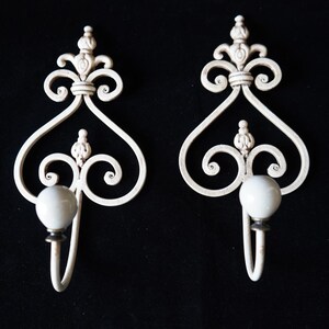 May include: Two ornate, off-white wall sconces with a scrolled design and a heart-shaped silhouette. Each sconce features a white, spherical knob and a hook. The background is solid black.