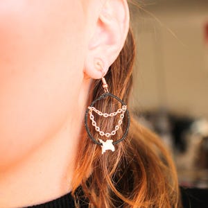 May include: A close-up of a black and gold hoop earring with a chain design and a small gold charm. The earring is hanging from an ear. The earring has a black hoop with gold chain and a gold charm.