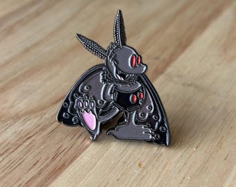 May include: A dark gray enamel pin featuring a moth-like creature holding a goblet with red accents. The pin has a pink paw and intricate wing details. The moth has large eyes and a smiling expression.