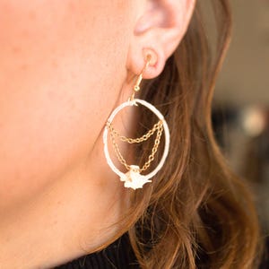 May include: Gold-tone hoop earrings with a white textured outer ring and dangling gold chains. A small, white floral accent hangs from the chains. The earrings are designed to be worn on the ear.