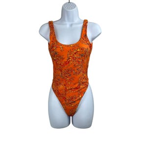 May include: Orange one-piece swimsuit with a flame pattern in red, yellow, and black. The swimsuit has a scoop neckline and thick straps. The high-cut leg openings and back are visible.