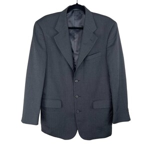 May include: A dark gray blazer with a three-button closure, two front pockets, and a chest pocket. The blazer is hanging on a black hanger against a white background. The jacket has a notched lapel and a subtle striped pattern.