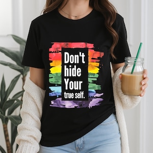Könnte beinhalten: Schwarzes T-Shirt mit dem Text "Don't hide Your true self." Der weiße Text ist vor einem Regenbogenhintergrund platziert. Das T-Shirt hat einen Rundhalsausschnitt.
