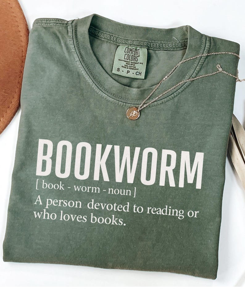 May include: An olive green t-shirt with the word "BOOKWORM" printed in white. The definition of bookworm is printed below the word. A gold necklace with a pendant rests on the shirt. The shirt is folded.