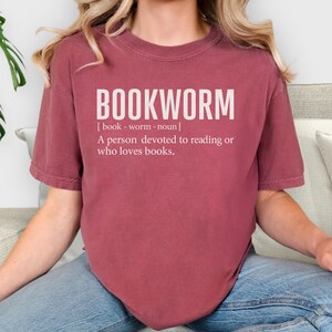 May include: A maroon t-shirt with the word "BOOKWORM" in large white letters. Below the word is a definition: "A person devoted to reading or who loves books."