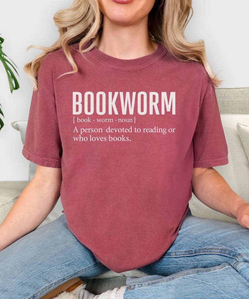May include: A maroon t-shirt with the word "BOOKWORM" in large white letters. Below the word is a definition: "A person devoted to reading or who loves books."