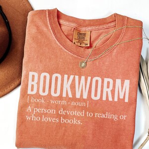 May include: Coral-colored t-shirt with the word "BOOKWORM" in large white letters. The definition of bookworm is printed below. A gold necklace with a small pendant rests on the shirt. A brown hat is partially visible in the upper left corner.