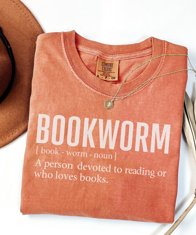 May include: Coral-colored t-shirt with the word "BOOKWORM" in large white letters. The definition of bookworm is printed below. A gold necklace with a small pendant rests on the shirt. A brown hat is partially visible in the upper left corner.