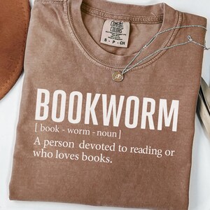 May include: A brown t-shirt with the word "BOOKWORM" in large white letters. The definition of bookworm is printed below the word. A silver necklace with a small pendant rests on the shirt.
