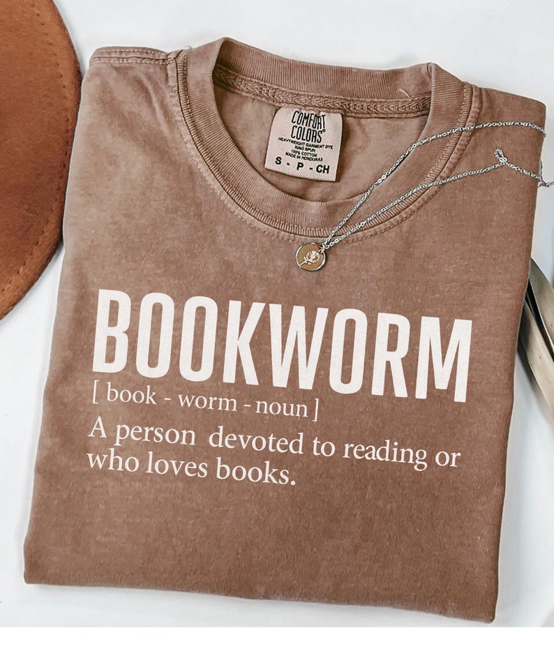 May include: A brown t-shirt with the word "BOOKWORM" in large white letters. The definition of bookworm is printed below the word. A silver necklace with a small pendant rests on the shirt.