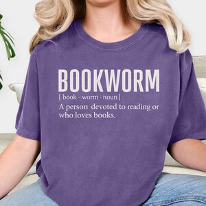 May include: A purple t-shirt with the word "BOOKWORM" in large white letters. Below the word is the definition: "A person devoted to reading or who loves books."