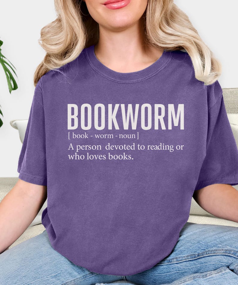 May include: A purple t-shirt with the word "BOOKWORM" in large white letters. Below the word is the definition: "A person devoted to reading or who loves books."