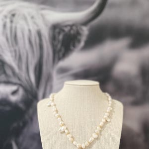 May include: A pearl necklace with a gold-coloured pendant in the shape of the letter "M" displayed on a mannequin. The necklace has a delicate design with small pearls and gold accents. The background is a black and white image of a Highland cow.