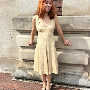 May include: A light beige sleeveless dress with a scoop neckline and a flared skirt. The dress has vertical seams and is worn with gold sandals. The background features a brick wall and a stone structure.