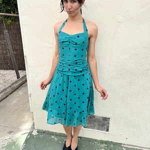 Op de afbeelding: Een blauwgroene halterjurk met een donkerblauw stippenpatroon, een geplooid lijfje, een gerimpelde taille en een rok tot op de knie met een ruchezoom. De jurk wordt gecombineerd met zwarte sandalen met open teen en gekruiste bandjes.