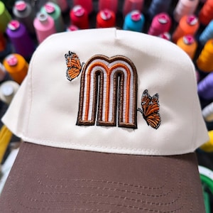 Puede incluir: Una gorra de béisbol color crema con visera marrón. El panel frontal presenta una letra m minúscula bordada en marrón, naranja y blanco, flanqueada por dos diseños de mariposas monarca naranjas y negras a los lados izquierdo y derecho.