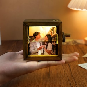 Könnte beinhalten: Kleine Musikbox aus Holz mit rechteckigem Rahmen. Die Box zeigt ein Foto eines Paares, das von warmem Licht beleuchtet wird. Eine Kurbel ist an der Seite sichtbar. Die Box wird in einer Hand gehalten.