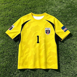Pode incluir: Uma camisola desportiva de manga curta em amarelo brilhante com um padr&atilde;o de camuflagem tonal subtil, pain&eacute;is laterais pretos e o n&uacute;mero 1 impresso a preto no peito. O emblema da FCF est&aacute; no peito esquerdo e um emblema na manga direita.