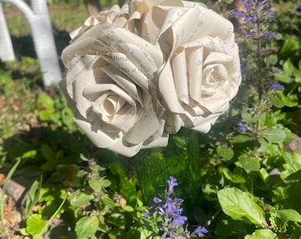 Puede incluir: Un ramo de rosas de papel hechas con las páginas de un libro, dispuestas en un jarrón de vidrio verde. Las rosas son blancas y el jarrón tiene una superficie texturizada. El ramo está sobre un lecho de hojas verdes.
