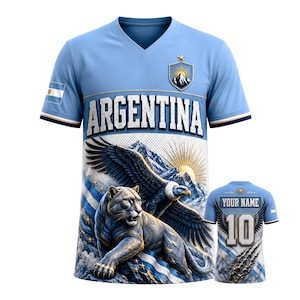 May include: A light blue V-neck t-shirt featuring the word ARGENTINA in white letters, a mountain crest, an eagle, and a puma. The sleeve displays the Argentina flag. The back shows the number 10 and the text YOUR NAME with eagle wings.