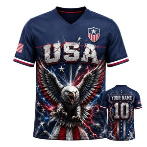 Può includere: Una t-shirt blu navy con scollo a V raffigurante un'aquila, fulmini rossi e blu e la scritta USA in un font effetto pietra crepata. Le maniche presentano strisce rosse e bianche e una bandiera americana sulla manica destra. Il retro mostra il numero 10 e il testo YOUR NAME.