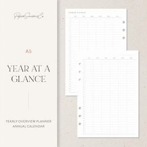 Può includere: Un pianificatore annuale A5 con il testo "Year at a Glance". Il pianificatore ha uno sfondo bianco con un design minimalista. Le pagine sono preformattate con sezioni mensili e giornaliere per la pianificazione del calendario annuale. Il logo Paper Seasons Co. si trova nell'angolo in alto a sinistra.