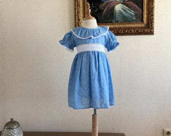 Puede incluir: Un vestido azul claro con cuello y cintura de encaje blanco. El vestido tiene mangas cortas y una falda acampanada, exhibido en un maniquí de madera. El vestido está hecho de una tela ligera con un estampado de lunares.