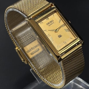 May include: A gold-toned Seiko Quartz watch with a rectangular face and mesh band. The watch face displays Roman numerals and the Seiko logo. The band is marked "Stainless Steel".
